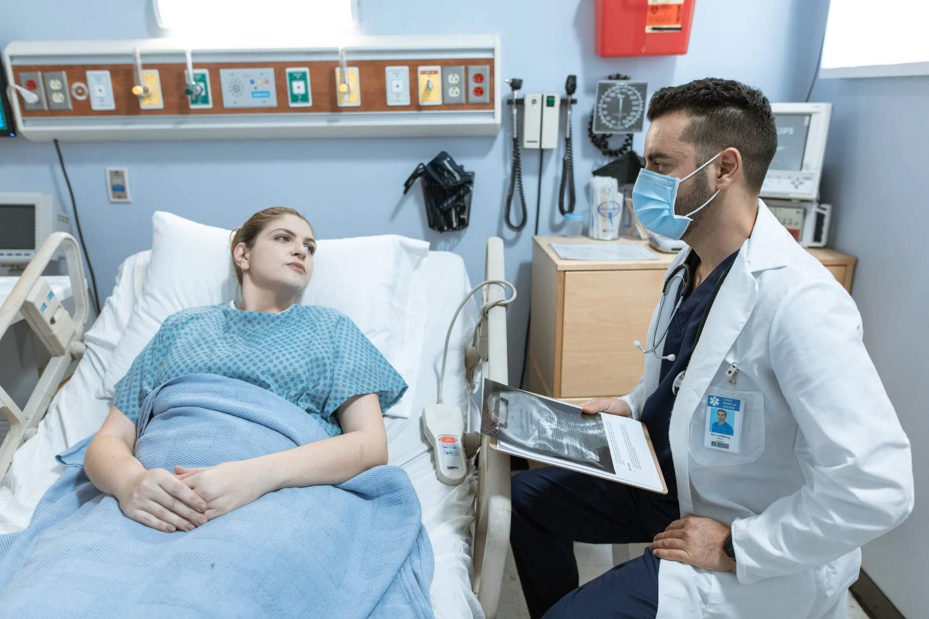 A clinician holding a clipboard with a profile image of a head and neck image, sitting at the bedside of a female patient in a hospital bed.