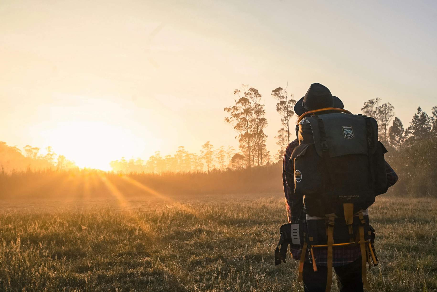 Hiker watching the sunrise in a forest clearing, wearing a large backpack and outdoor gear.